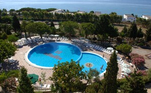 A large circular swimming pool surrounded by lounge chairs and umbrellas, with a view of the sea and buildings in the background.