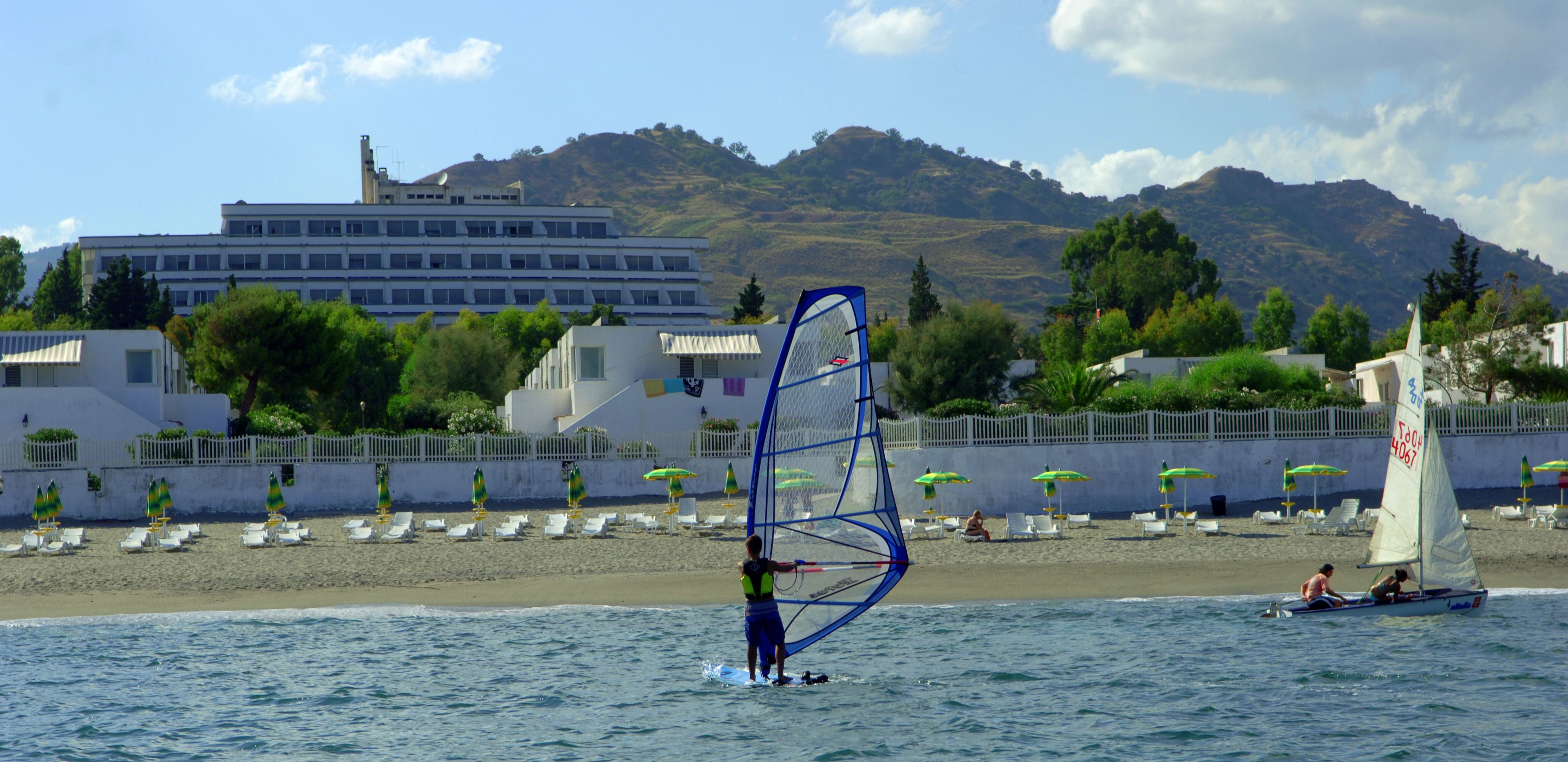 Une plage où l’on pratique la planche à voile, un bâtiment en arrière-plan et un paysage montagneux.