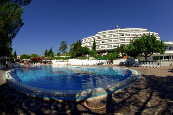 A large outdoor swimming pool with lounge chairs and umbrellas, surrounded by trees and a modern building in the background.