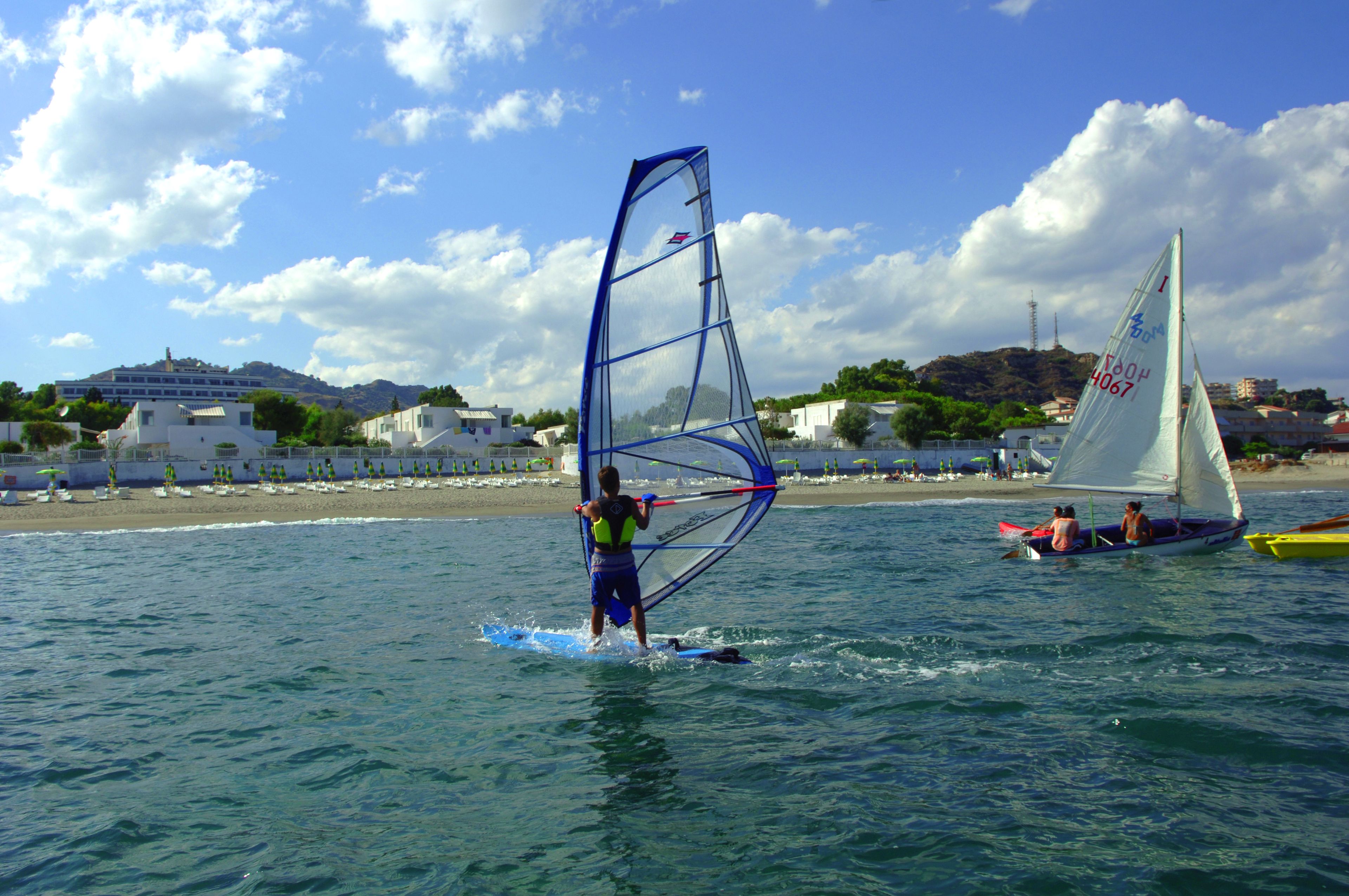 A beach scene with a person windsurfing, a sailboat, and buildings in the background.