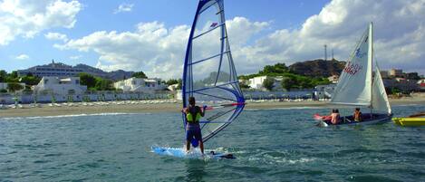 A beach scene with a person windsurfing, a sailboat, and buildings in the background.