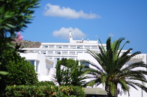 A white building with multiple balconies and a satellite dish, surrounded by greenery and palm trees.