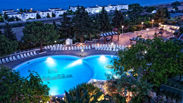 A swimming pool with lounge chairs and a sunbathing area, surrounded by trees and buildings with a view of the sea. - Hotel Villaggio Club ALTALIA (Brancaleone)