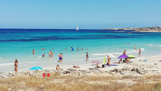Ein Strand mit klarem türkisblauem Wasser, Menschen beim Schwimmen und Sonnenbaden, und eine felsige Küstenlinie im Hintergrund.