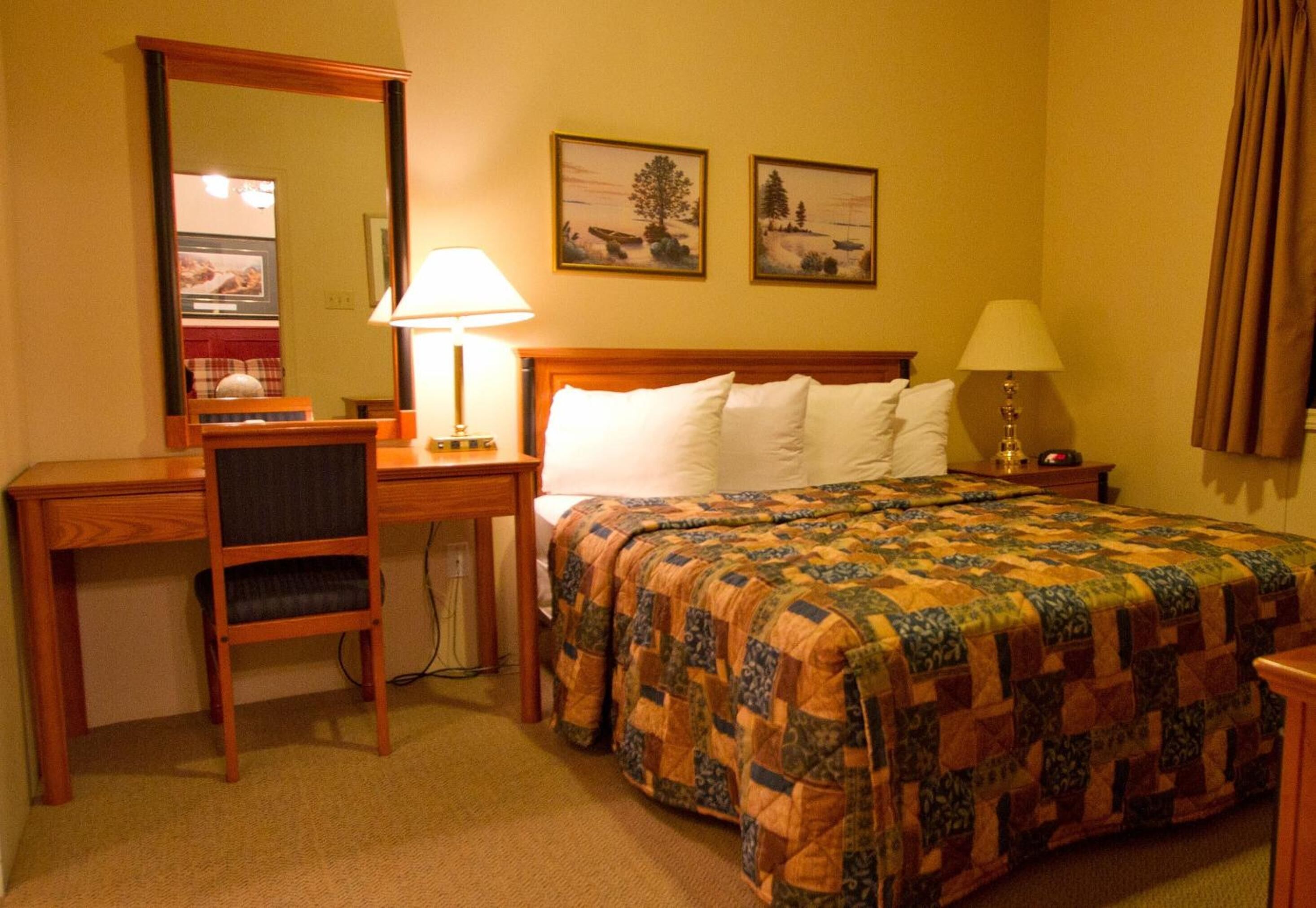 A hotel room with a wooden desk, a chair, a bed with a quilt, two wall-mounted lamps, and two framed pictures on the wall.