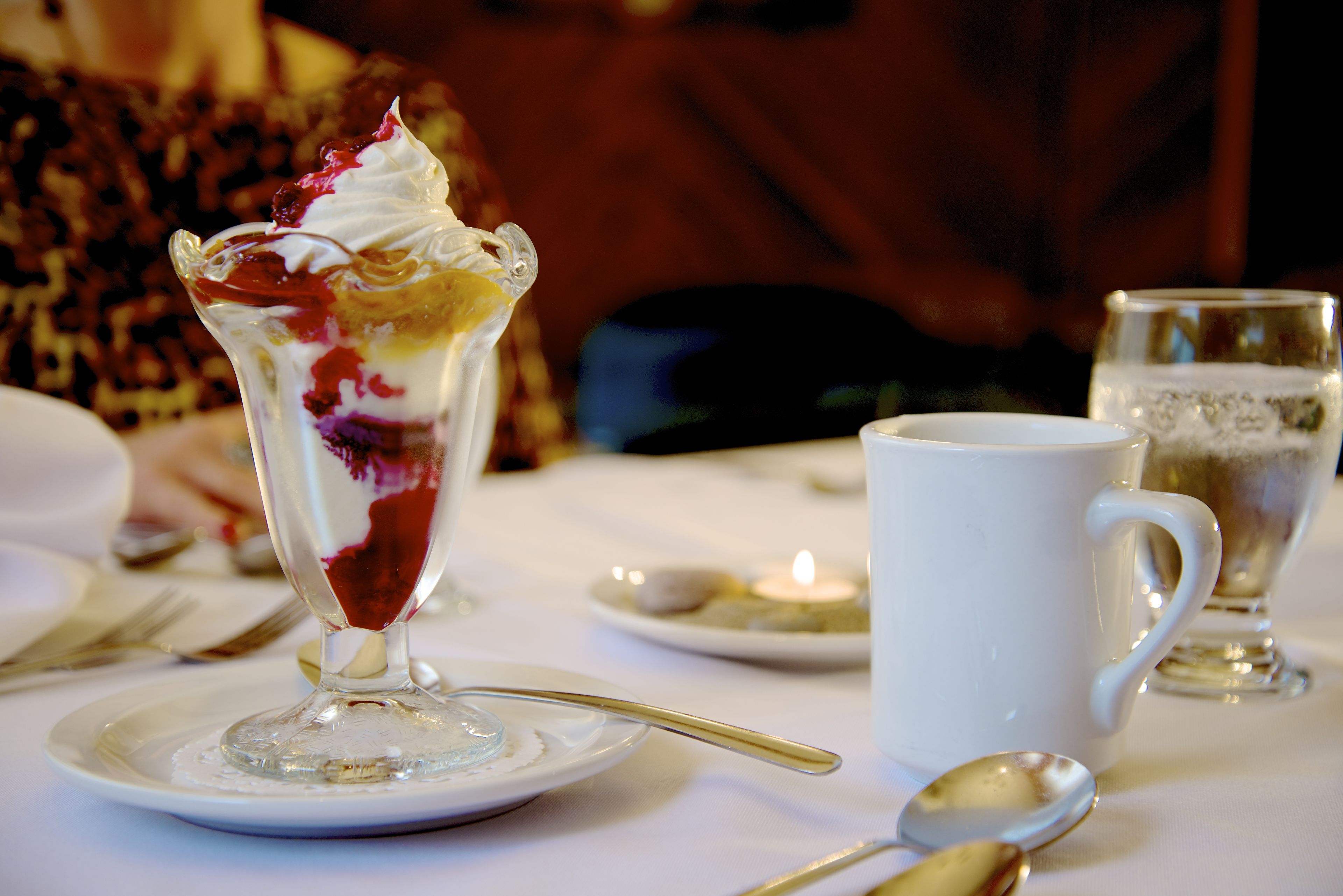 Un postre en un vaso sobre un plato blanco, una taza blanca y una cuchara sobre una mesa.
