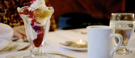Un postre en un vaso sobre un plato blanco, una taza blanca y una cuchara sobre una mesa.