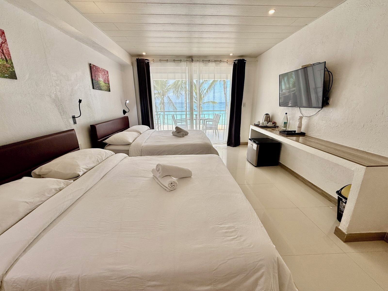 A hotel room with two beds, a TV, and a balcony with a view of palm trees and the sea.
