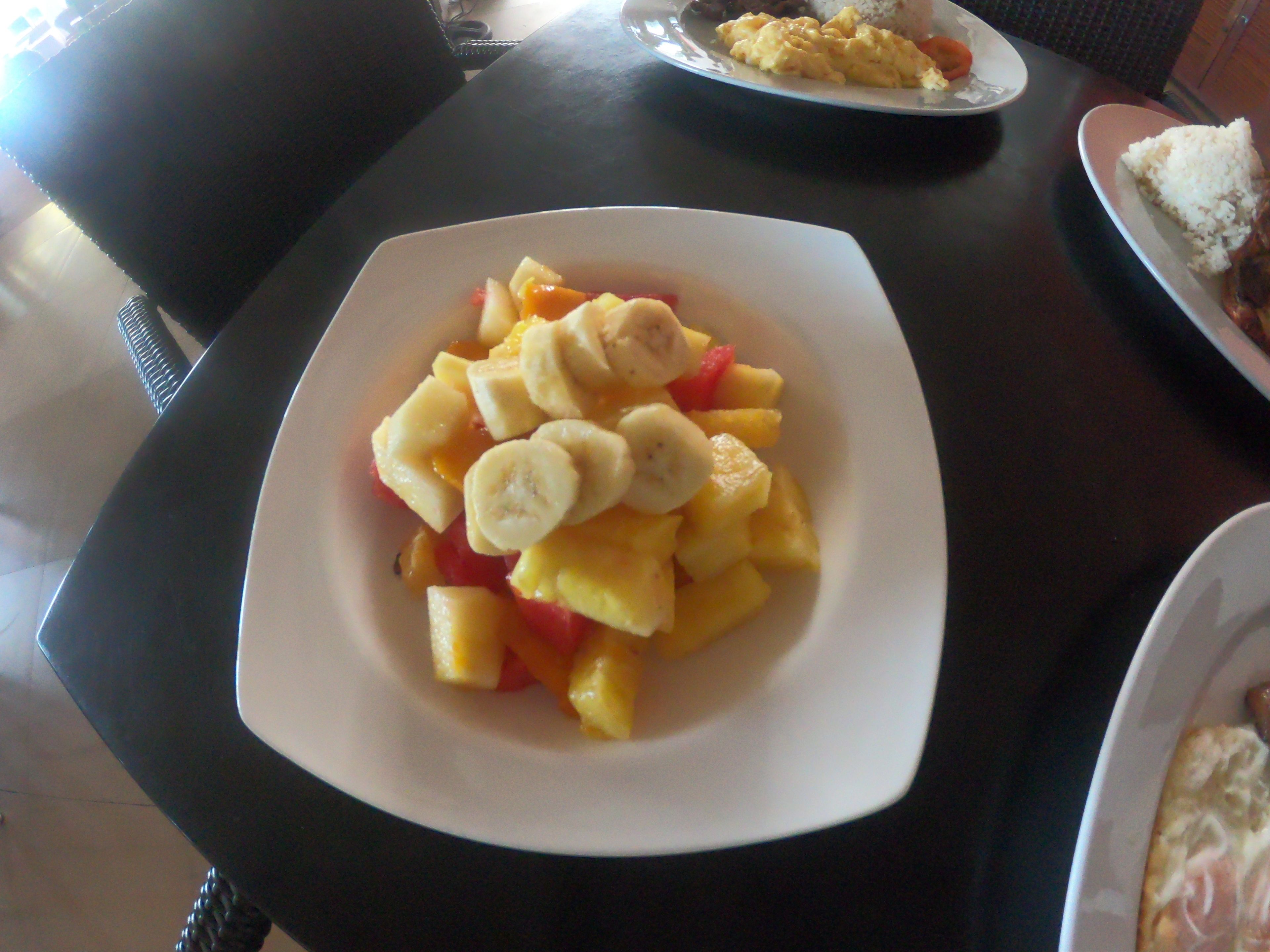 A dining table with a plate of fruit salad and a plate of scrambled eggs.