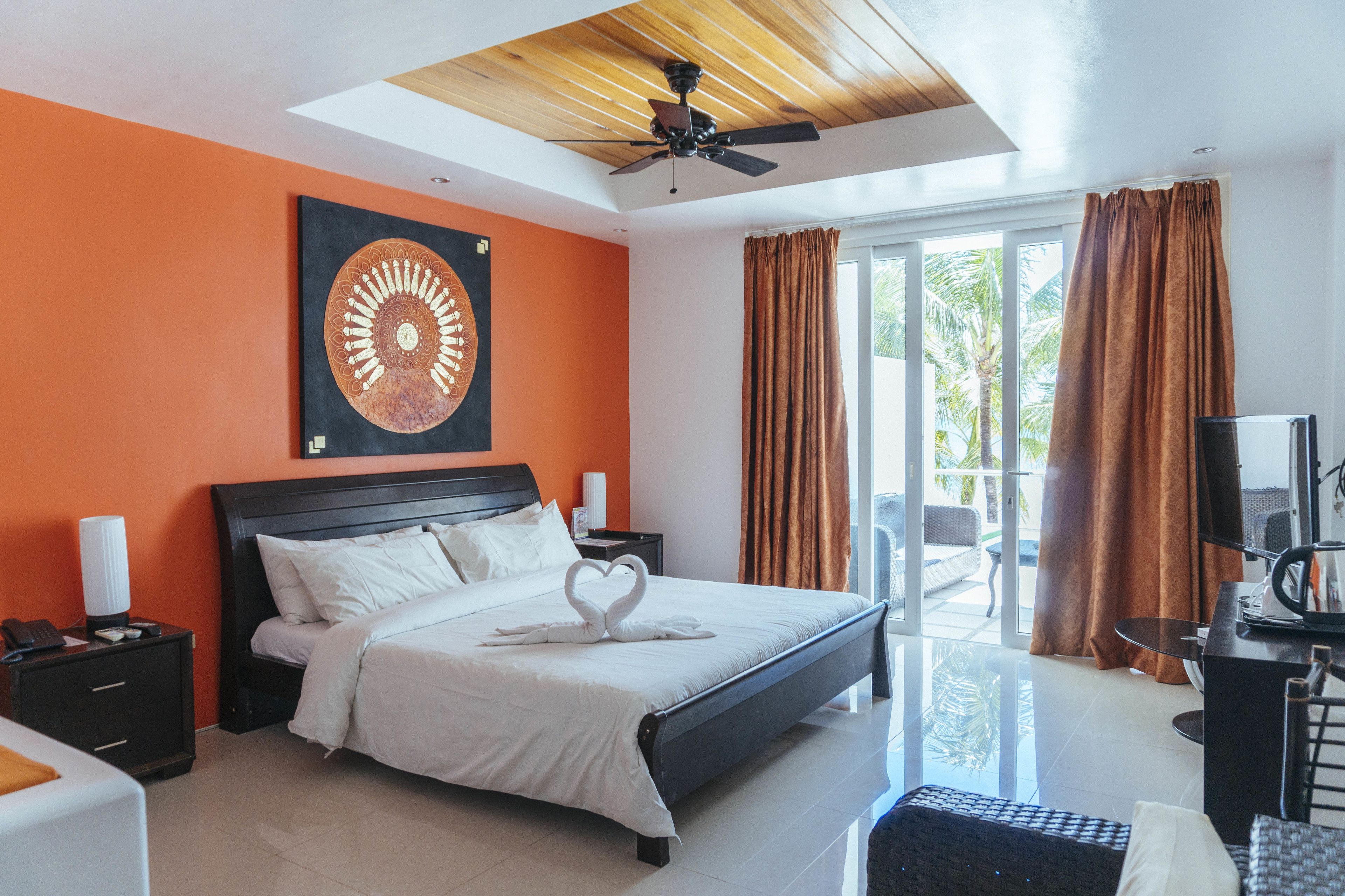 A hotel room with a large bed, a ceiling fan, a view of the outdoors, and a painting on the wall.