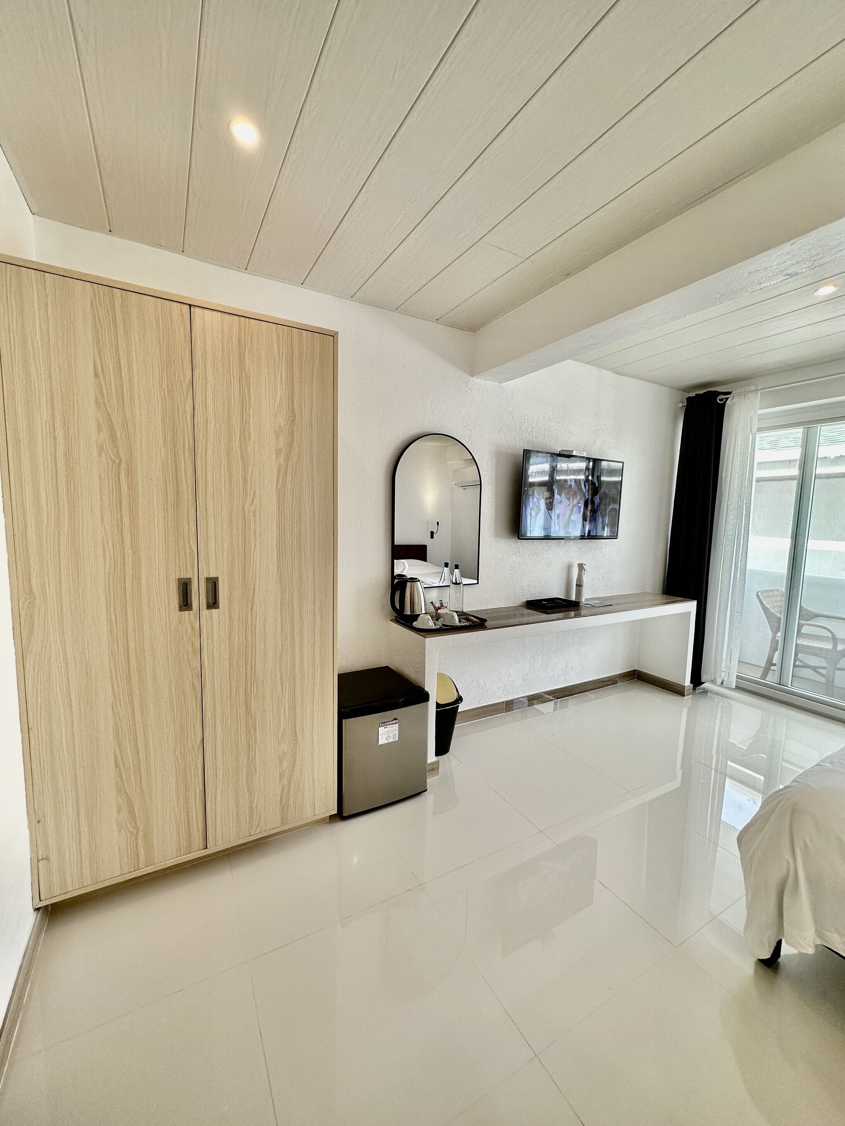 A modern hotel room with a wooden wardrobe, a small kitchenette, a flat-screen TV, and a large window offering a view.