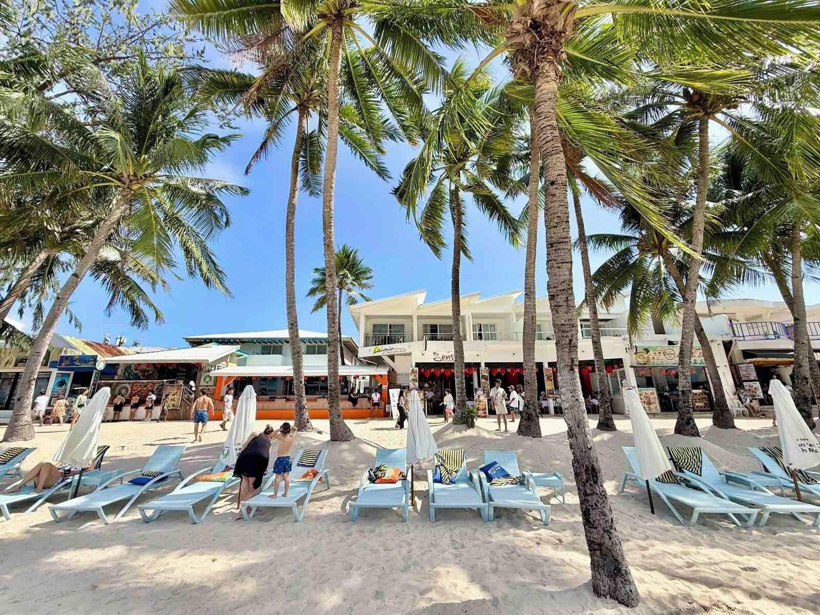Una playa con palmeras, tumbonas y un edificio con tiendas.