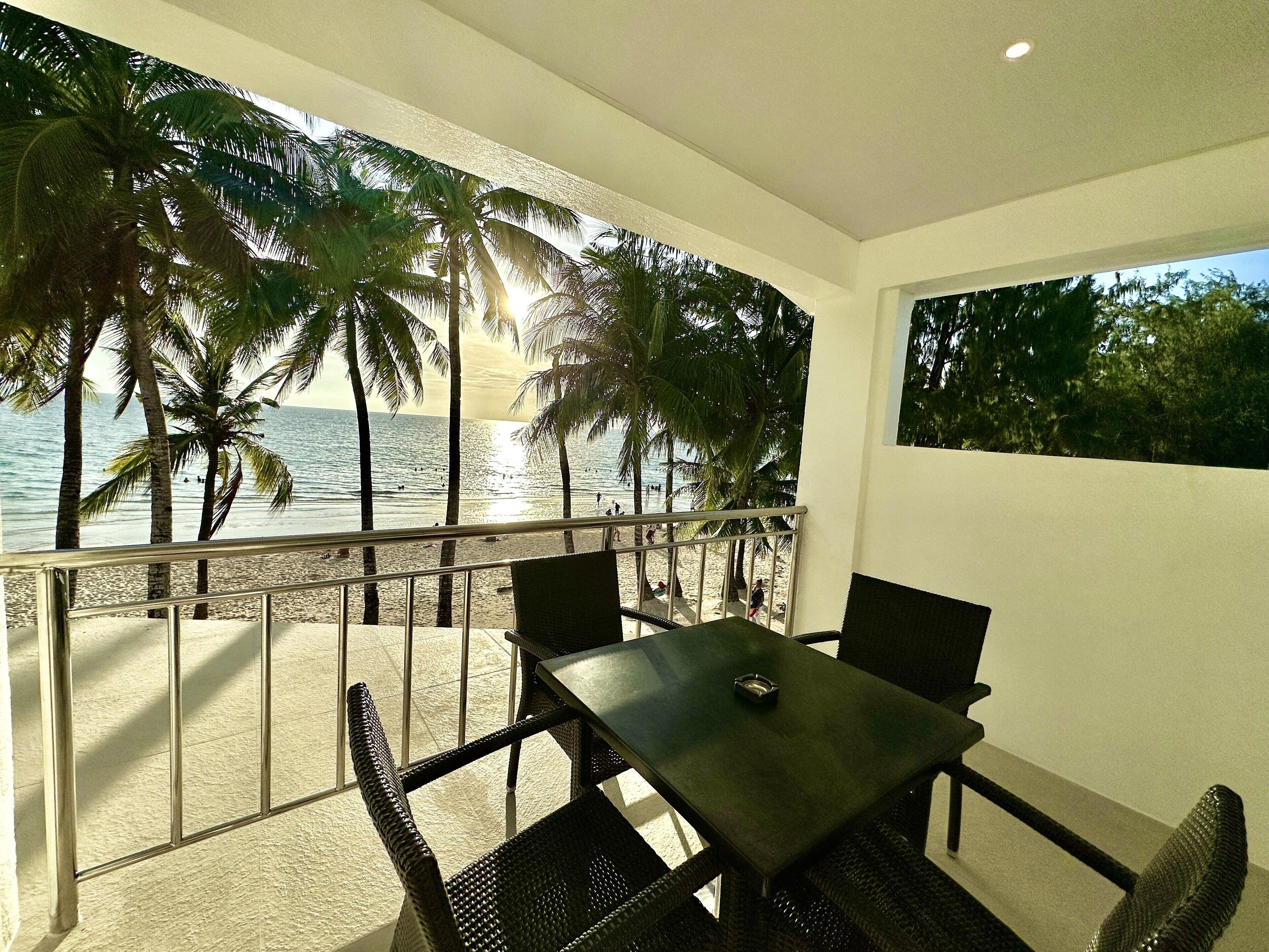 A balcony with a table and chairs overlooking a beach with palm trees and the ocean.