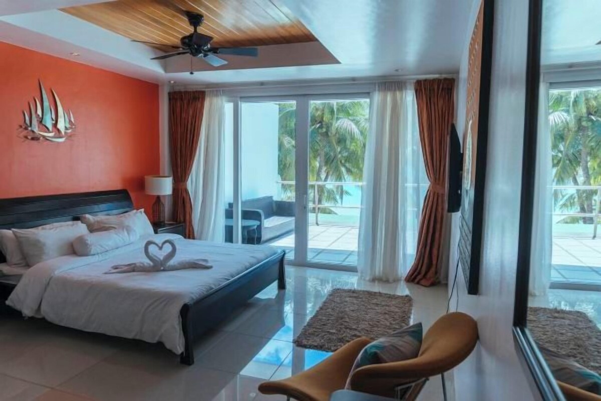 Une chambre d’hôtel avec un lit, un ventilateur de plafond, une grande fenêtre avec des rideaux et une vue sur des palmiers.