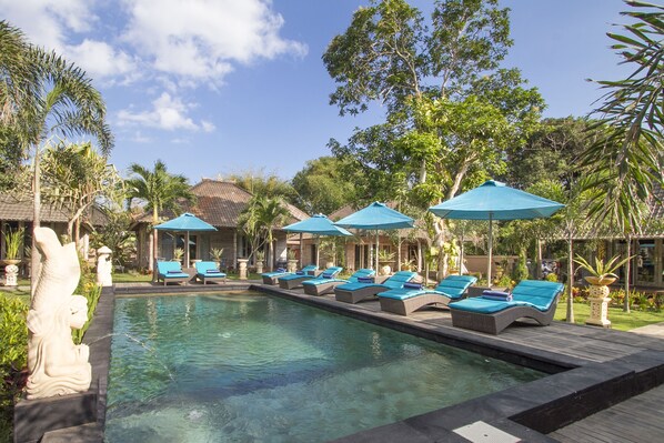 A pool area with lounge chairs and umbrellas, surrounded by tropical plants and a clear sky.