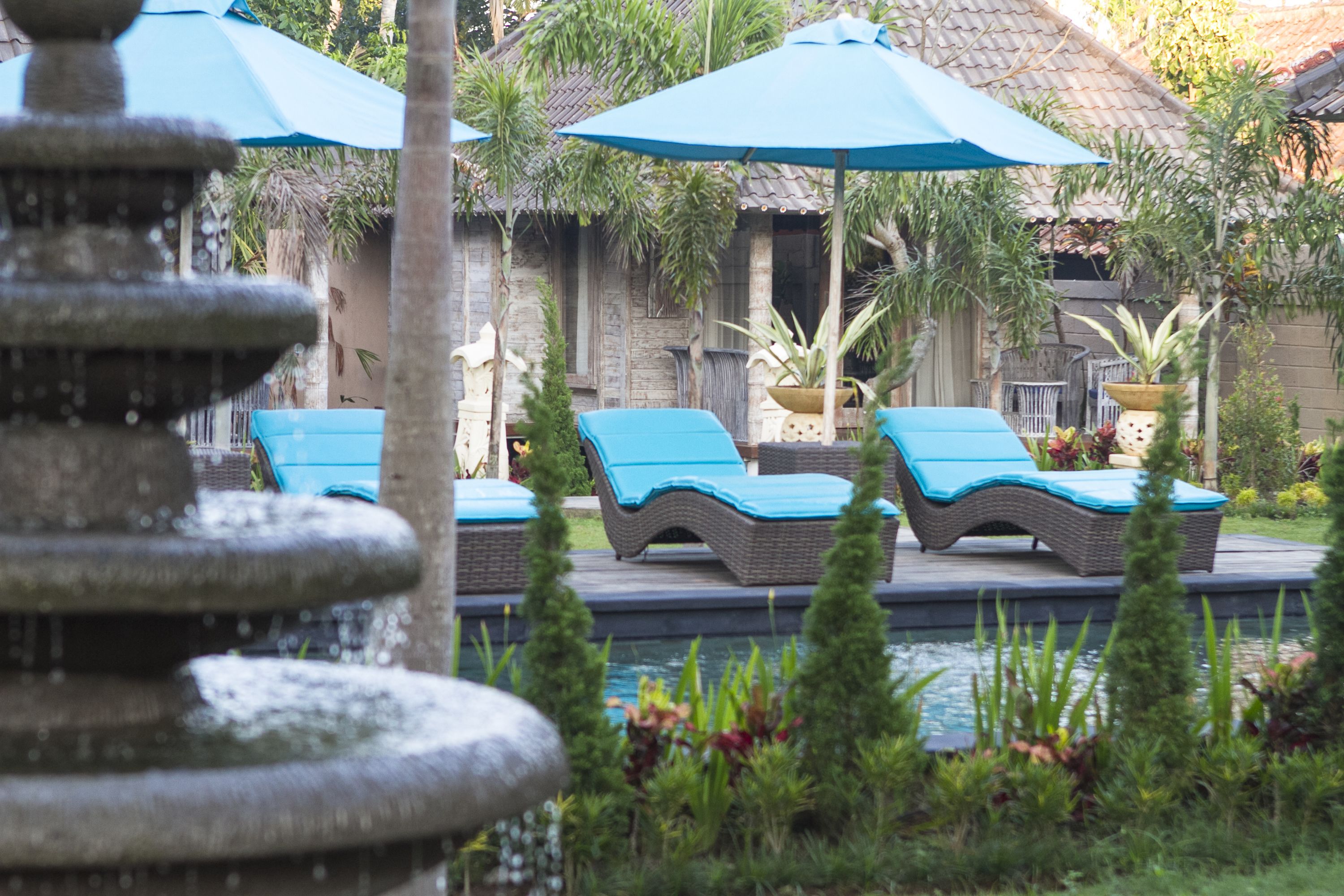 A pool area with lounge chairs and umbrellas, surrounded by a stone fountain and lush greenery.
