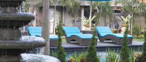 A pool area with lounge chairs and umbrellas, surrounded by a stone fountain and lush greenery.