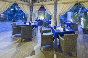 An outdoor dining area with wicker furniture and blue tablecloths, surrounded by a covered pergola and plants.