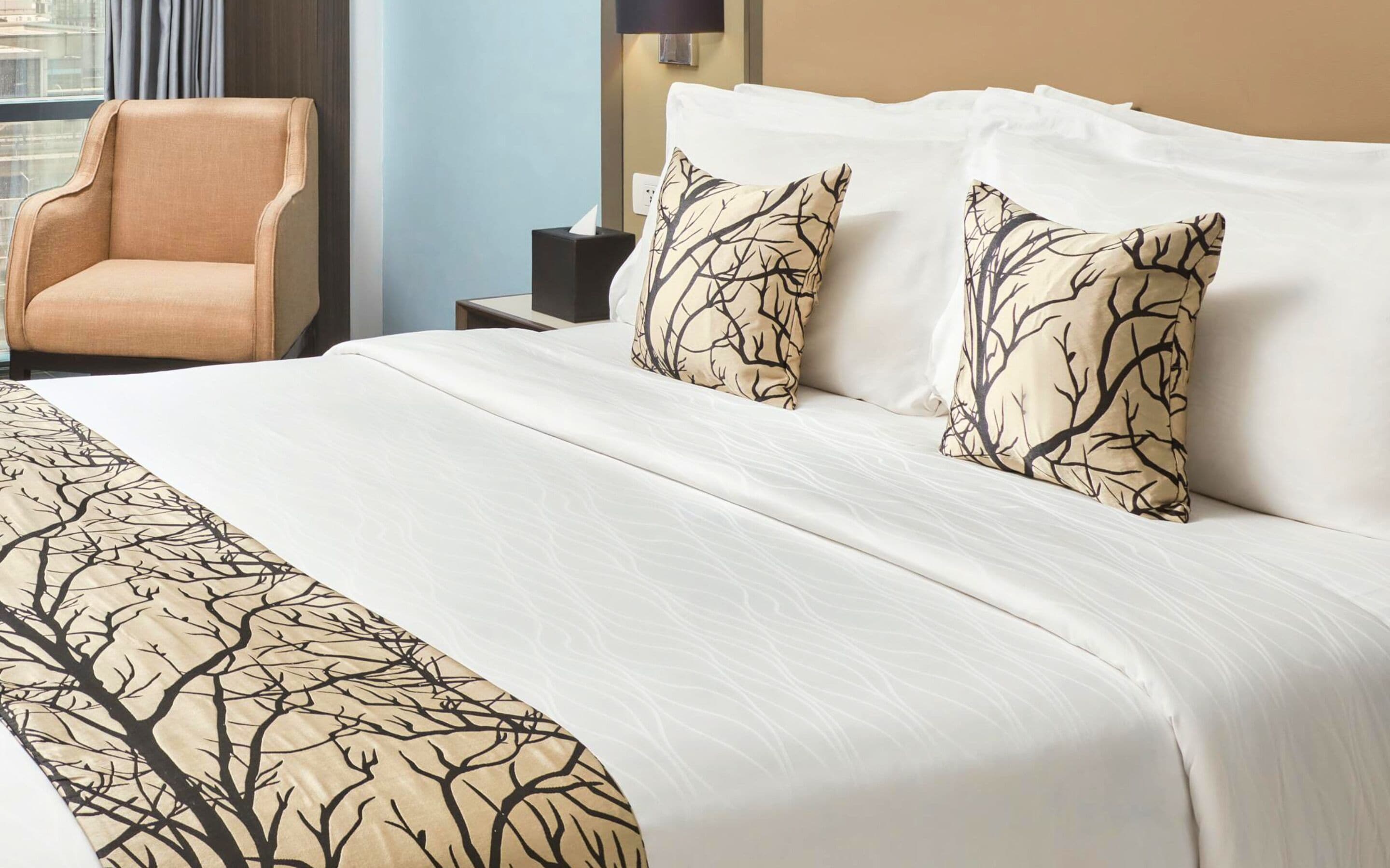 A hotel room with a large bed, two pillows with a tree pattern, a beige armchair, and a nightstand with a lamp.