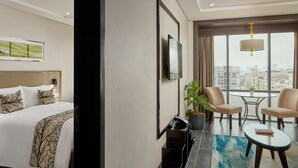 A hotel room with a bed, a TV, a potted plant, a seating area with chairs, and a view of the city through large windows. - Belmont Hotel Manila near NAIA Terminal 3 MNL (Pasay)