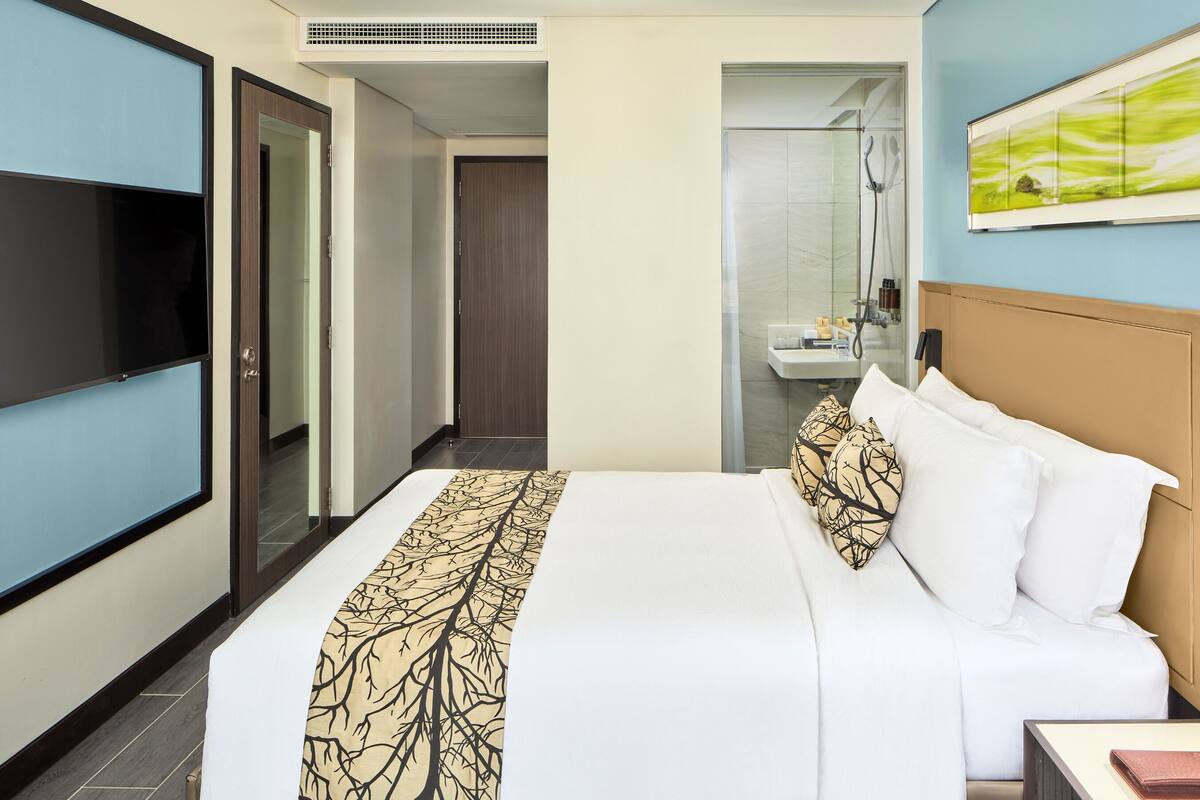 A hotel room with a bed, a TV, a bathroom with a shower, and a painting on the wall.