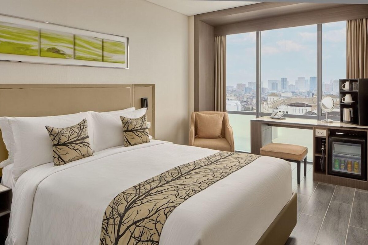 A hotel room with a large bed, a desk with a computer, a chair, and a view of the city.