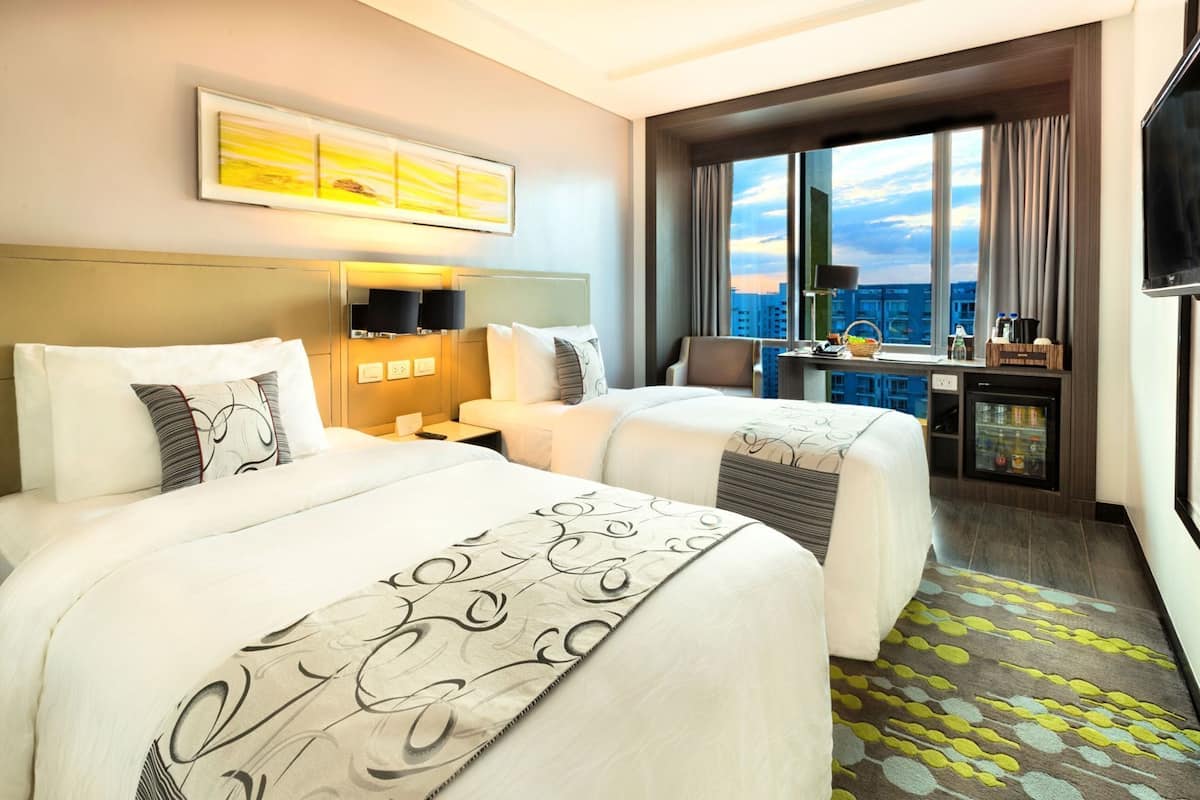A hotel room with two beds, a desk, a minibar, and a large window with a city view.