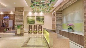 A hotel lobby with a reception desk, three chairs, a large abstract painting, a decorative ceiling chandelier, and a potted plant.