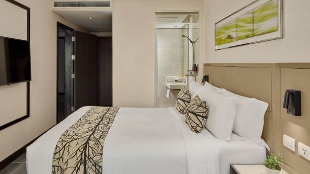 Una camera d'albergo con un letto, un comodino con una pianta, una televisione, un bagno parzialmente visibile attraverso una porta aperta e un quadro appeso al muro.