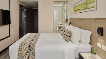Habitación de hotel con cama, mesita de noche con planta, televisor, baño visible a través de una puerta abierta y un cuadro en la pared.