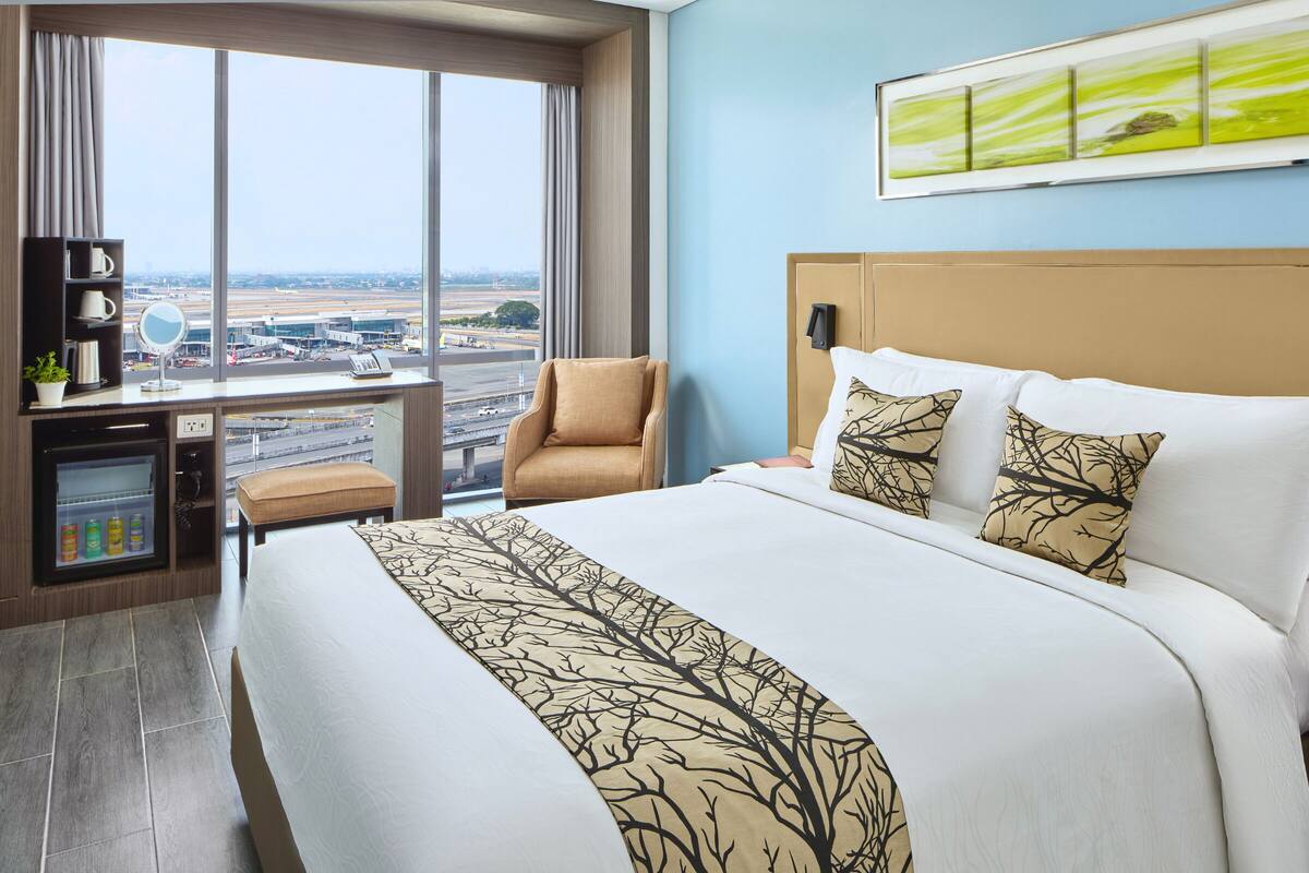 A hotel room with a large bed, a chair, a desk with a mirror, and a view of an airport.