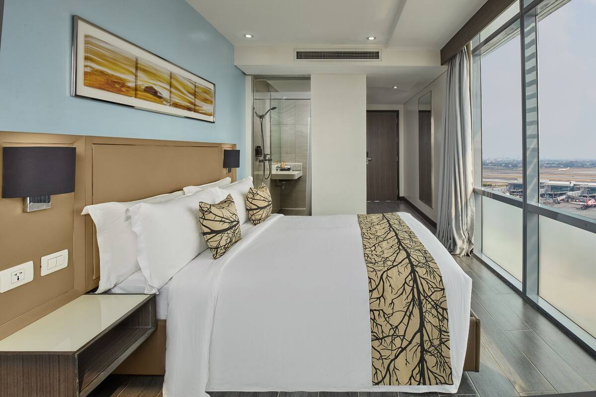 A hotel room with a large bed, a nightstand, a wall-mounted lamp, a bathroom with a glass shower, and a view of an airport.