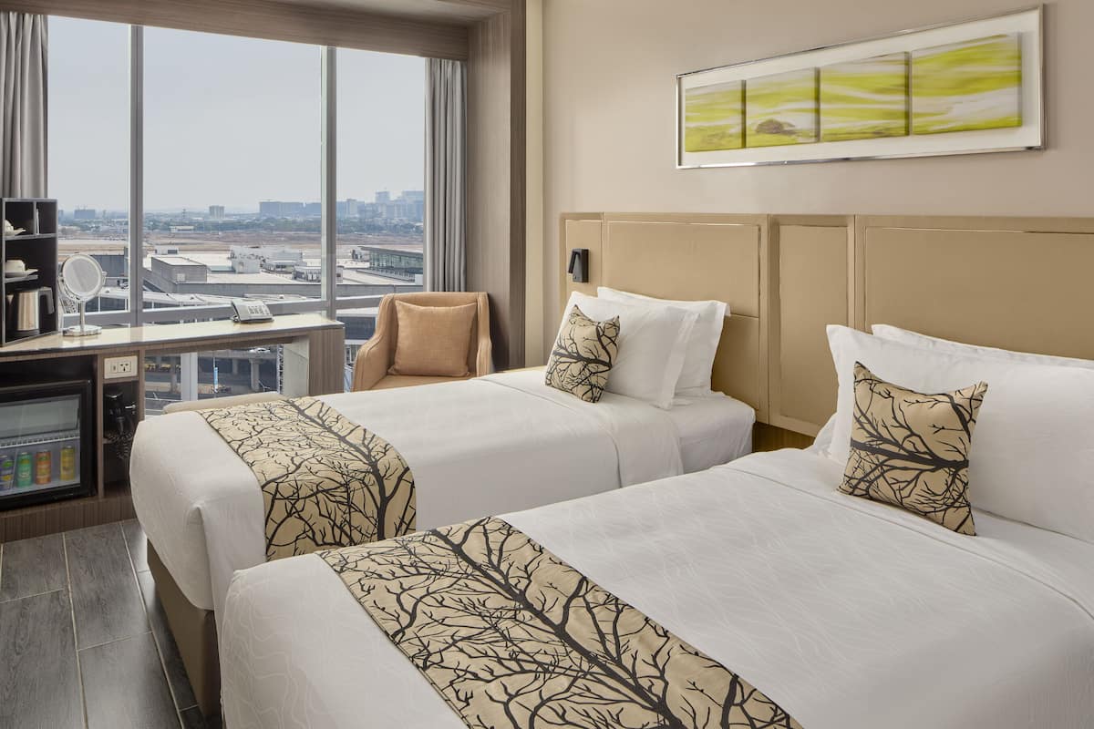 A hotel room with two beds, a desk with a computer, a mirror, and a view of the cityscape.