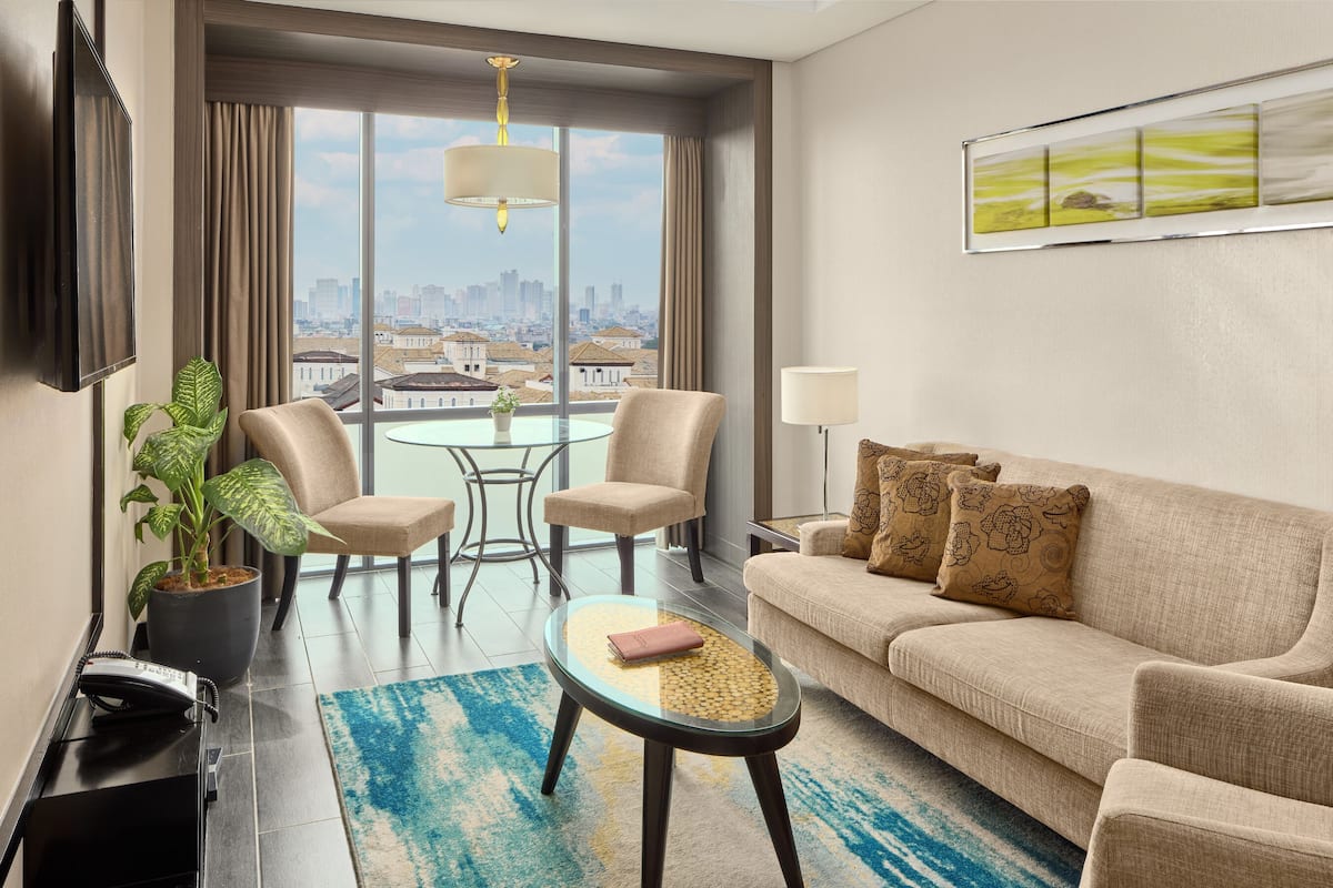 A modern hotel room with a city view, a sofa, two armchairs, a glass coffee table, a lamp, and a potted plant.