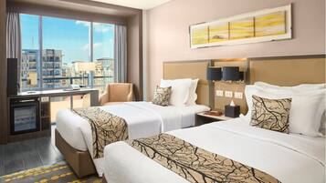 A hotel room with two beds, a desk, a chair, a TV, and a large window with a city view.