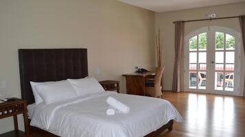 Una cama grande con sábanas blancas, un cabecero de madera y una mesita de noche con una lámpara. Hay un escritorio de madera con una silla y un teléfono. La habitación tiene un ventanal con cortinas y vistas a zonas verdes.