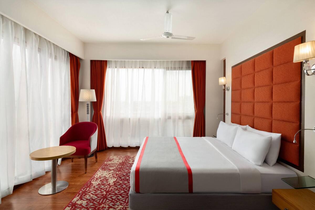 A hotel room with a bed, a round table, a red chair, a window with sheer curtains, and a ceiling fan.