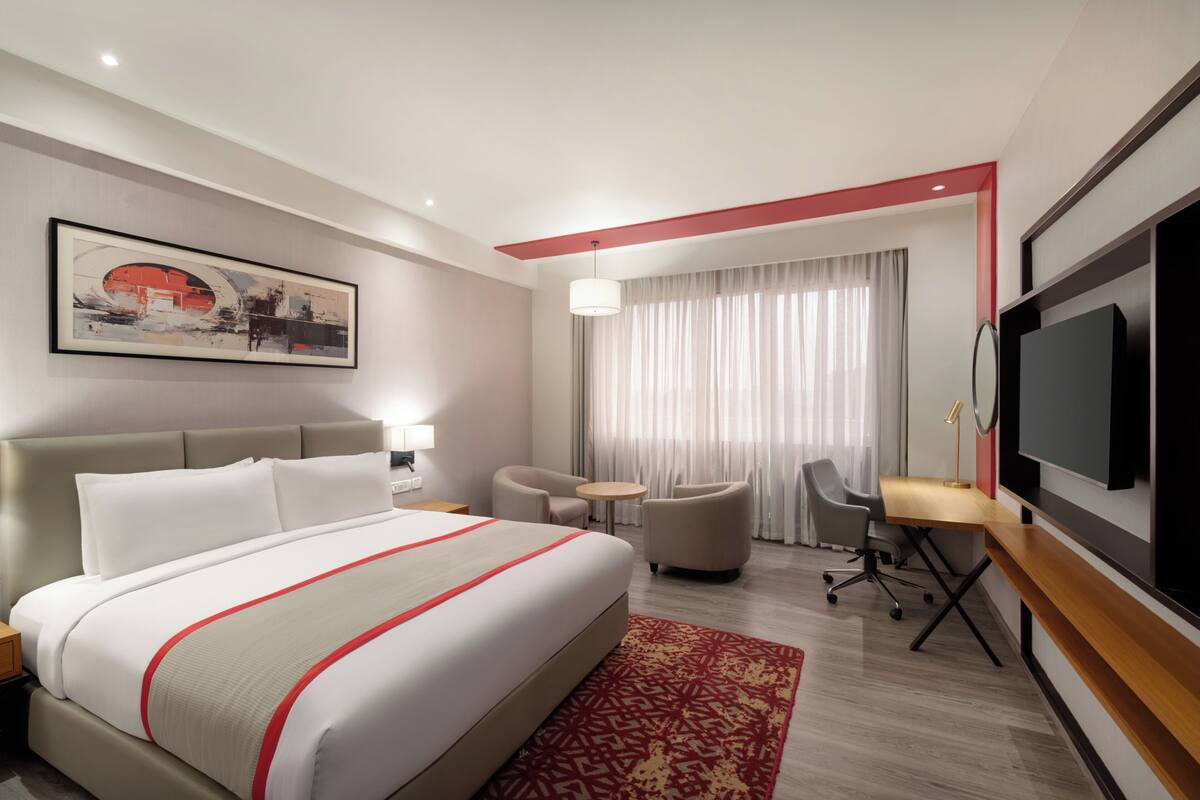 A hotel room with a large bed, a desk, two chairs, a TV, and a painting on the wall.