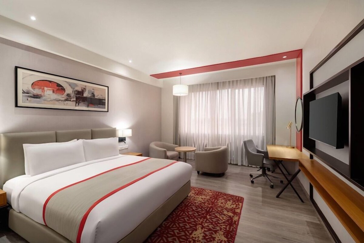 A hotel room with a bed, a desk, a chair, a television, and a painting on the wall.