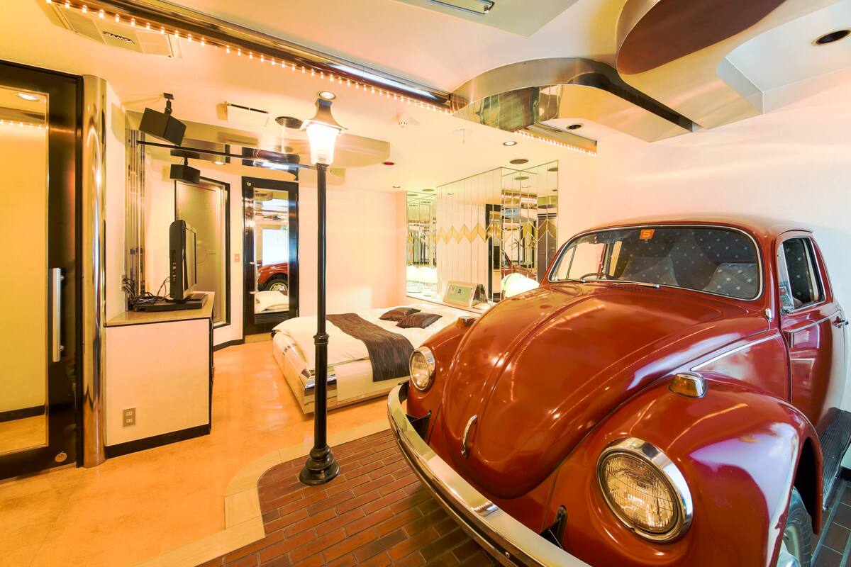 A vintage car in a hotel room with a bed, a desk, and a mirror.