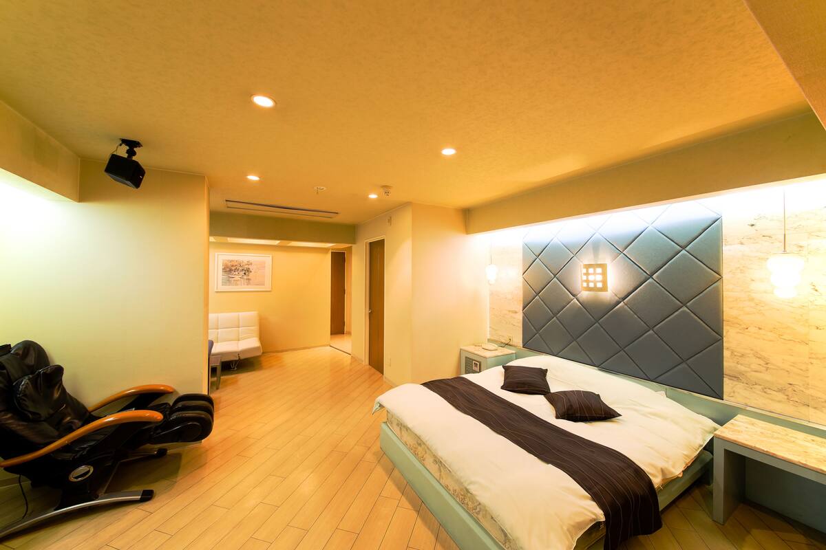 A hotel room with a bed, a massage chair, a bedside table, and a wall-mounted light fixture.