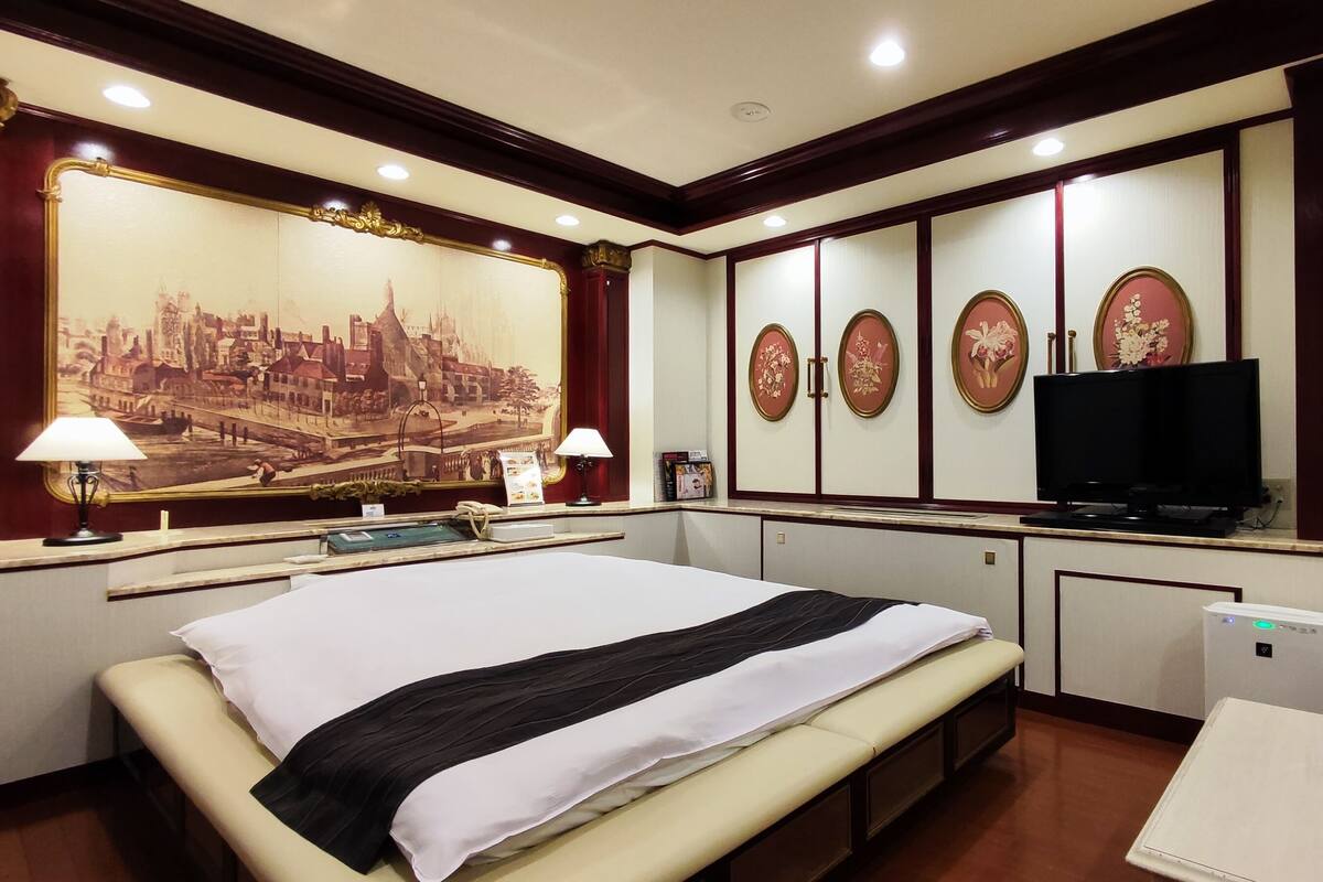 A hotel room with a large bed, a desk with a computer, a television, and a large cityscape painting on the wall.