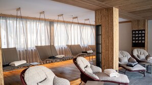A modern spa room with multiple lounge chairs, a wooden ceiling, and a large window with sheer curtains. - Hotel Post (Prutz)