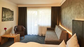 Ein Hotelzimmer mit einem großen Bett, einem Sofa, einem Sessel, einem kleinen Tisch und einem großen Fenster mit Vorhängen.
