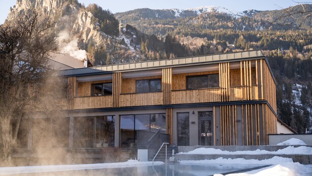 A modern building with a wooden facade, large windows, and a pool area with snow. - Hotel Post (Prutz)