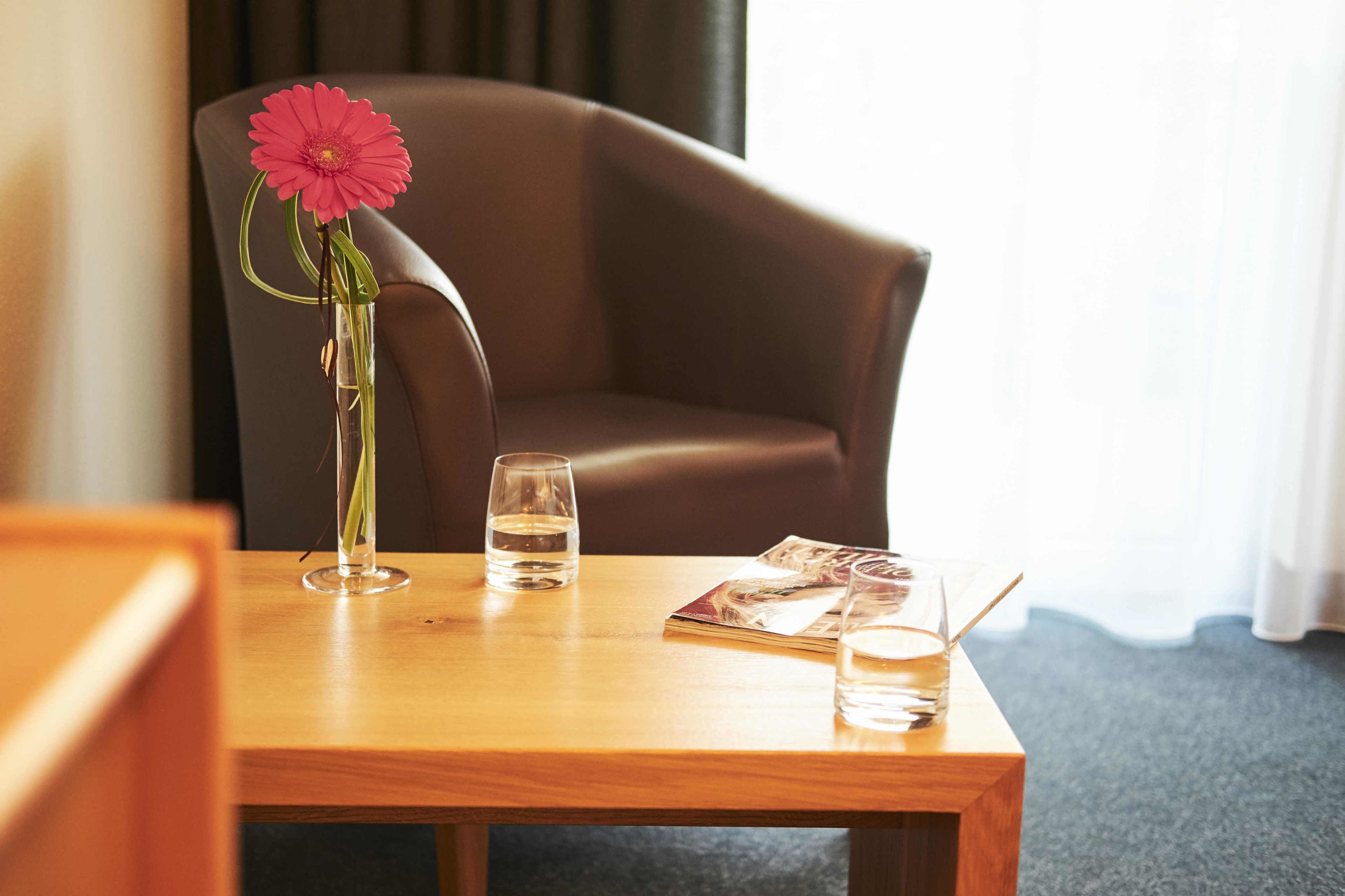 A wooden table with a vase of pink flowers, two glasses of water, and a magazine.