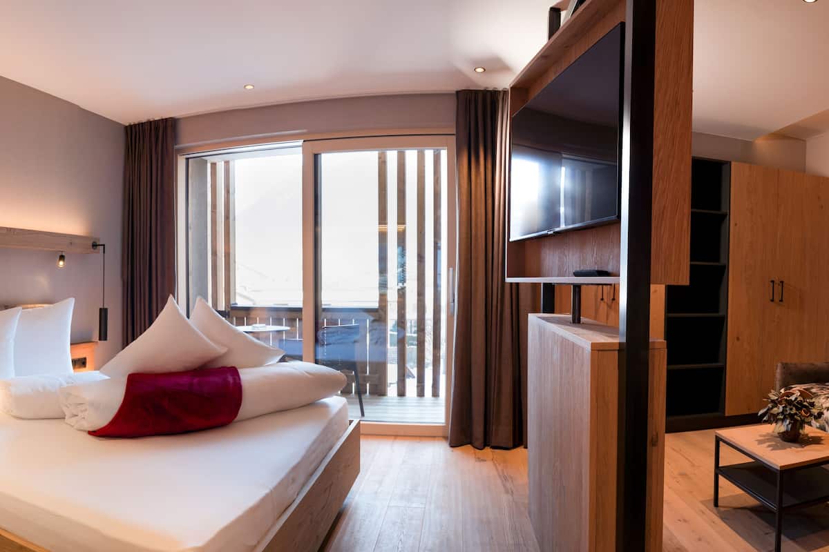 A modern hotel room with a large bed, a TV, a desk, and a balcony with curtains.