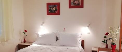Une chambre à coucher avec un lit, deux tables de chevet, une petite table, un vase rouge contenant une rose et deux tableaux encadrés accrochés au mur.