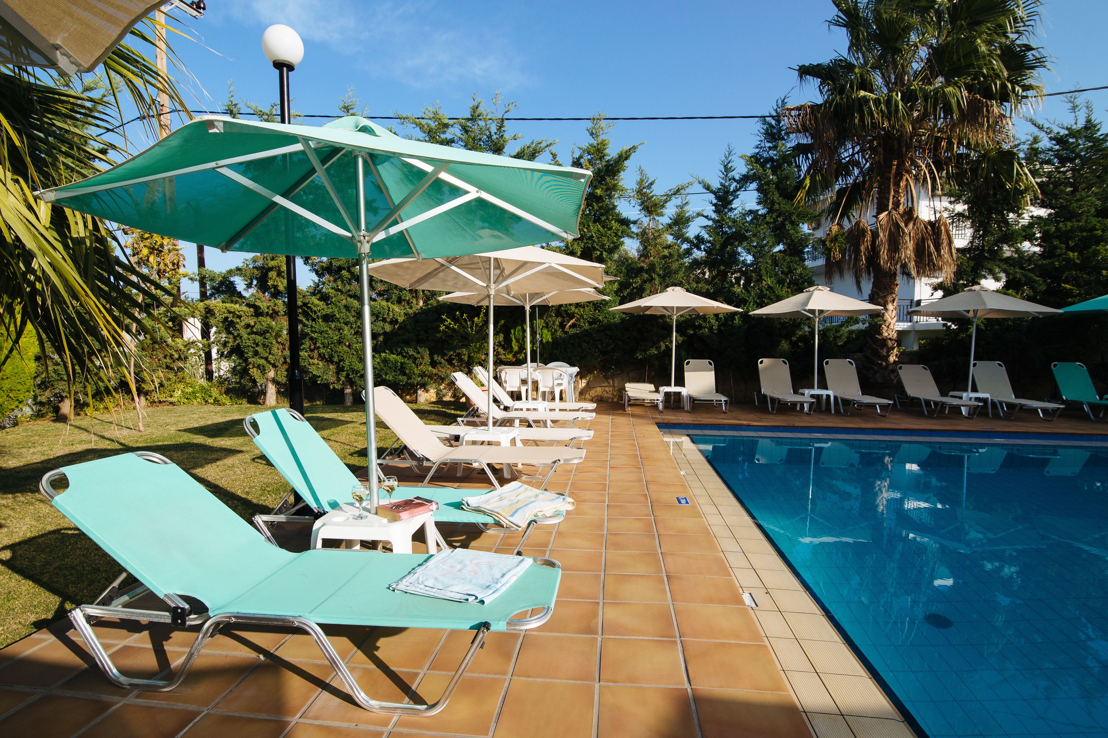 Un espace aménagé au bord de la piscine, avec des chaises longues et des parasols, une piscine et des palmiers en arrière-plan.