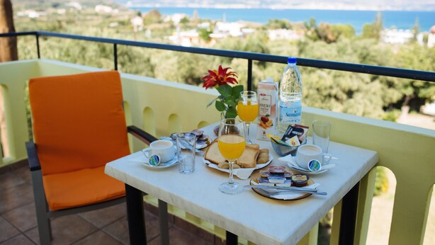 A balcony with a table set for breakfast, including toast, fruit, and beverages, overlooking a scenic landscape. - Kalithea Apartments (Apokoronas)